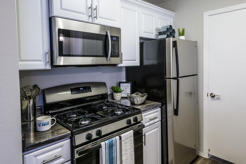 Renovated Kitchens - In Select Homes | Renovated kitchens featuring black fusion counter tops, wood-style flooring, and stainless steel appliances. We are excited to offer in-person tours while following social distancing and we encourage all visitors to wear a face covering.