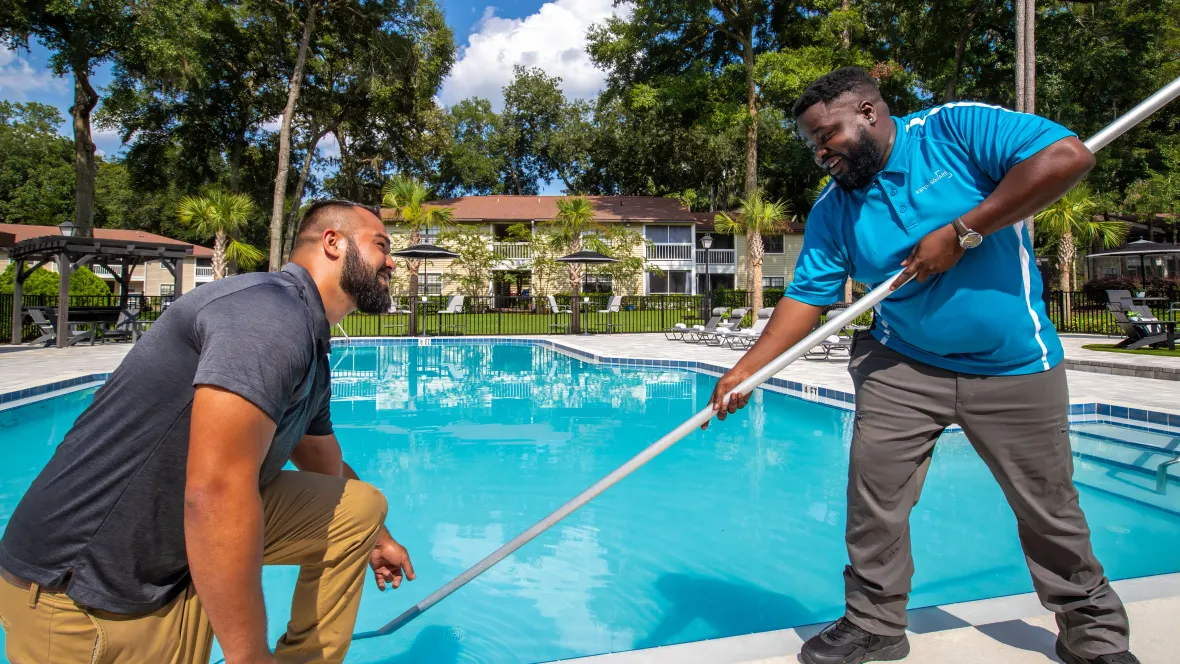 Two maintenance employees at the pool. One is skimming the pool and the other is kneeling down pointing at pool.