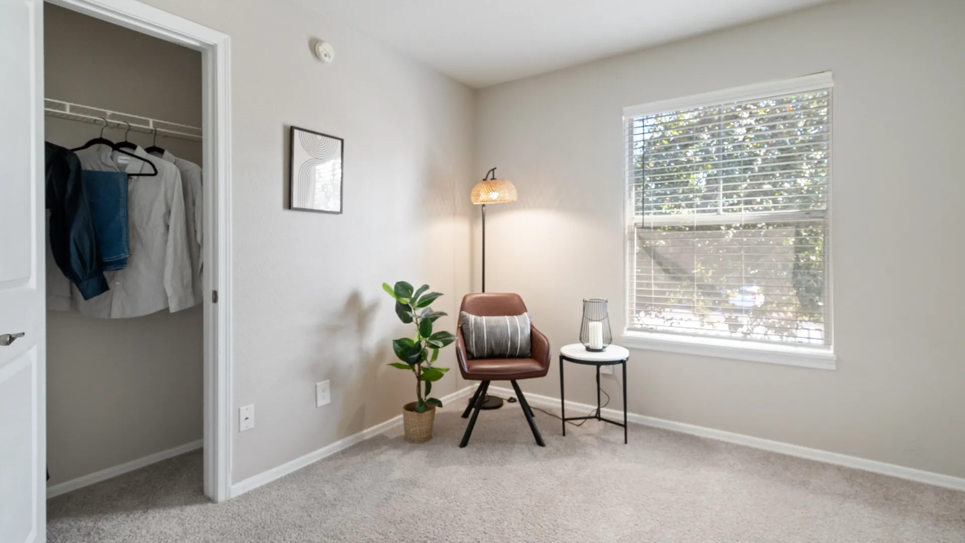 Well-lit bedroom with soft carpeting, a seating area, modern decor, and an open closet with hanging clothes.