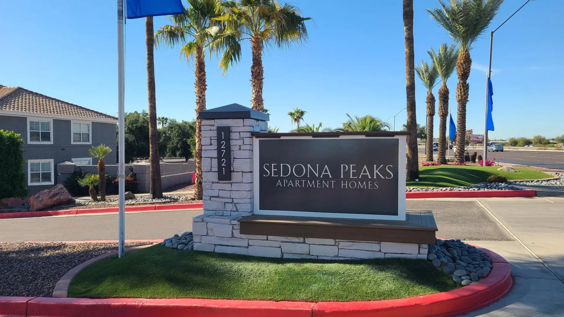 Entrance sign of Sedona Peaks Apartment Homes with a stone base, surrounded by palm trees, green landscaping, and a sunny sky in the background.