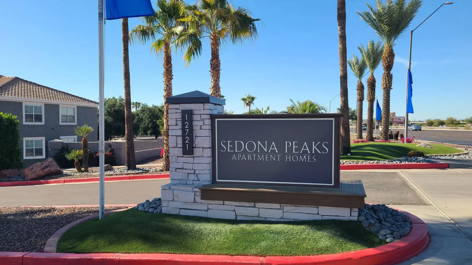 Entrance sign of Sedona Peaks Apartment Homes with a stone base, surrounded by palm trees, green landscaping, and a sunny sky in the background.