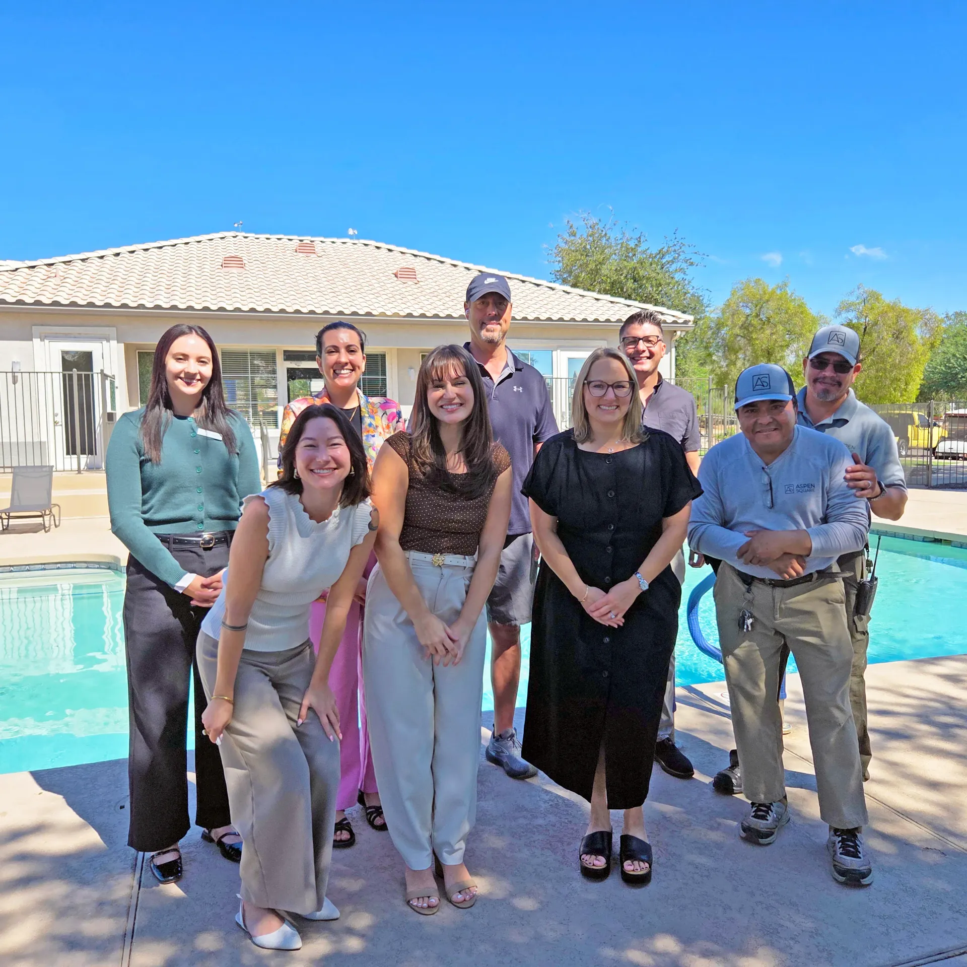 Nine Riverwood team members gathered poolside with the community swimming pool, clubhouse, and landscaped grounds in the background.