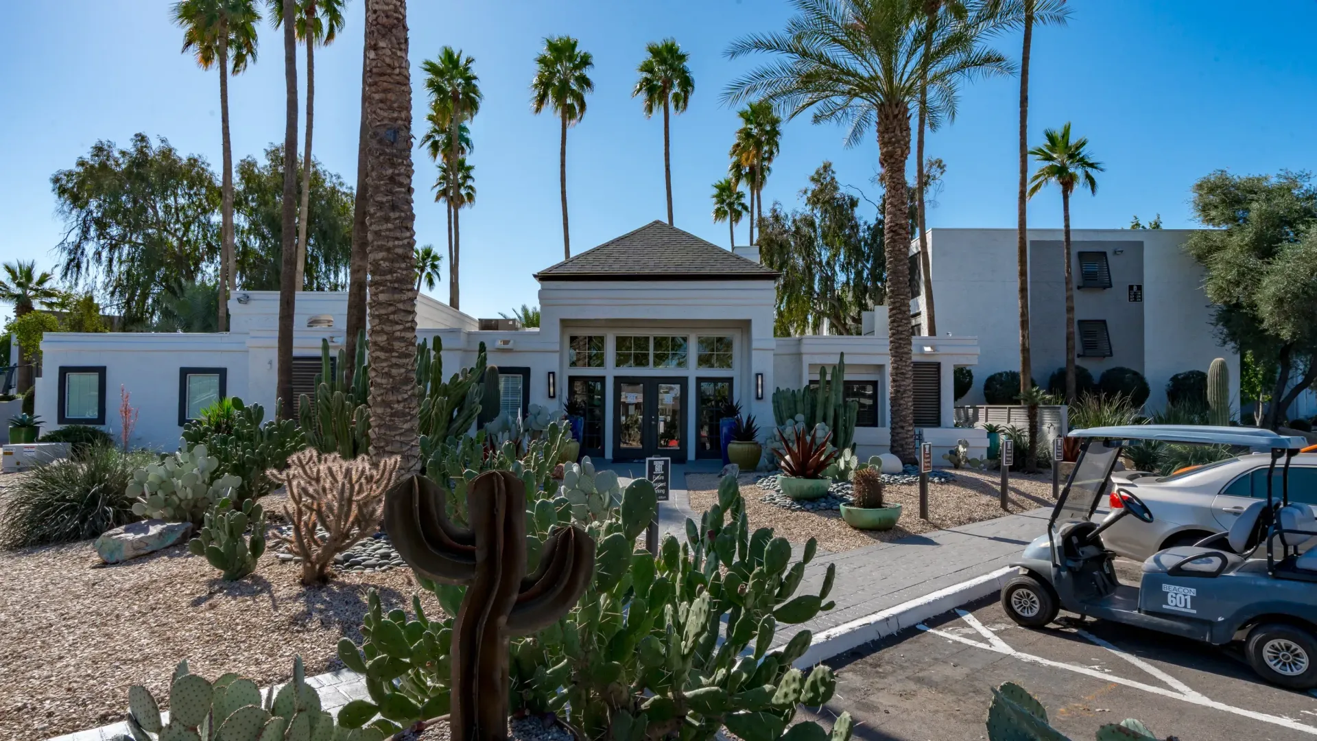 Sunlit path lined with cheerful yellow cacti and desert flora leads to the grand clubhouse entrance.