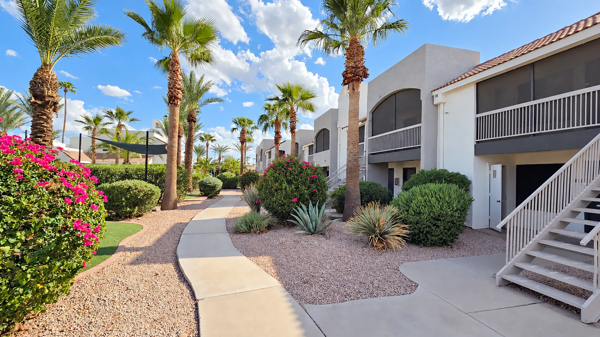Level 550 walkway winding past palm trees, pink flowering shrubs, desert landscaping, and apartment residences with screened balconies.