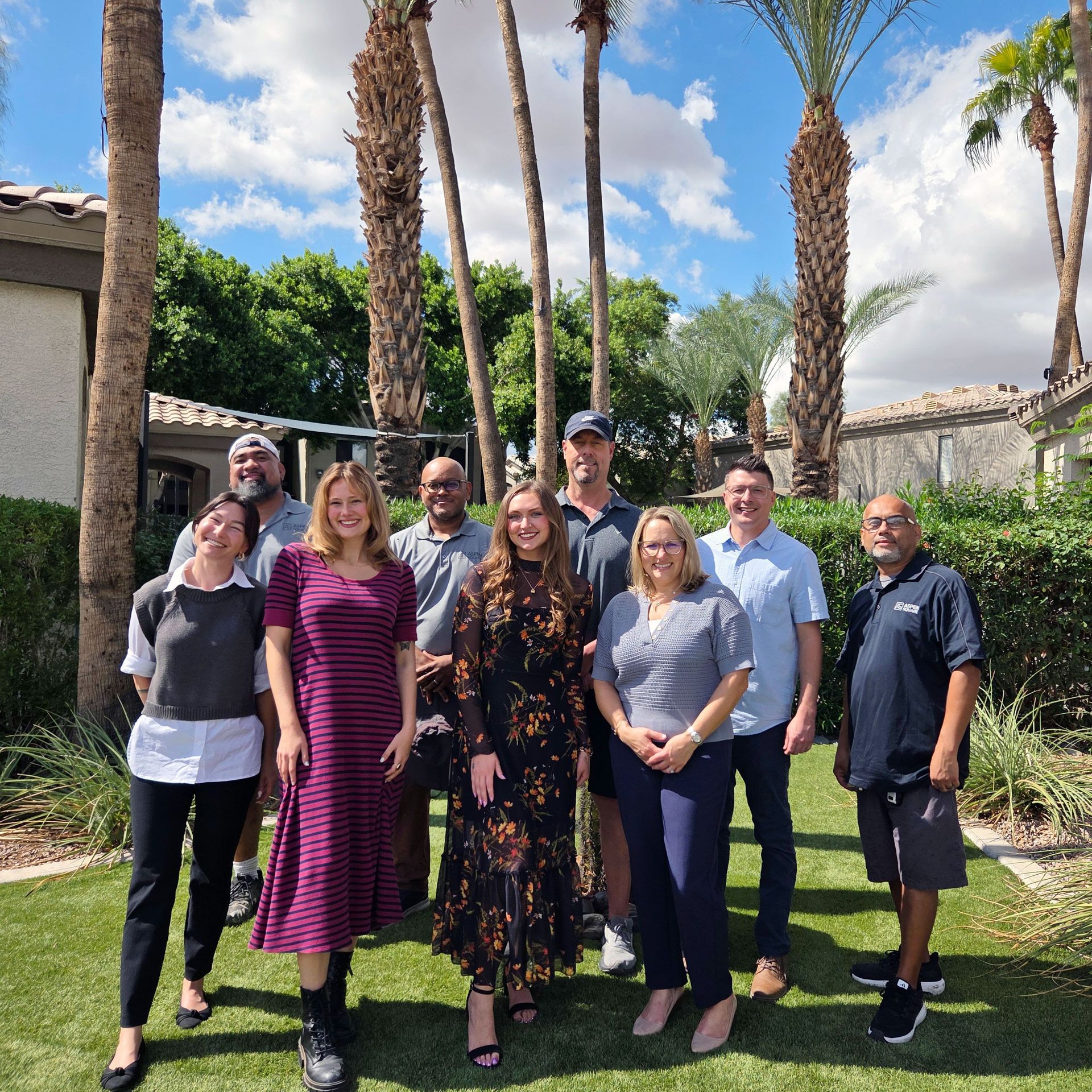 Nine Luxe 1930 team members photographed outdoors with palm trees, greenery, and community buildings in the background.