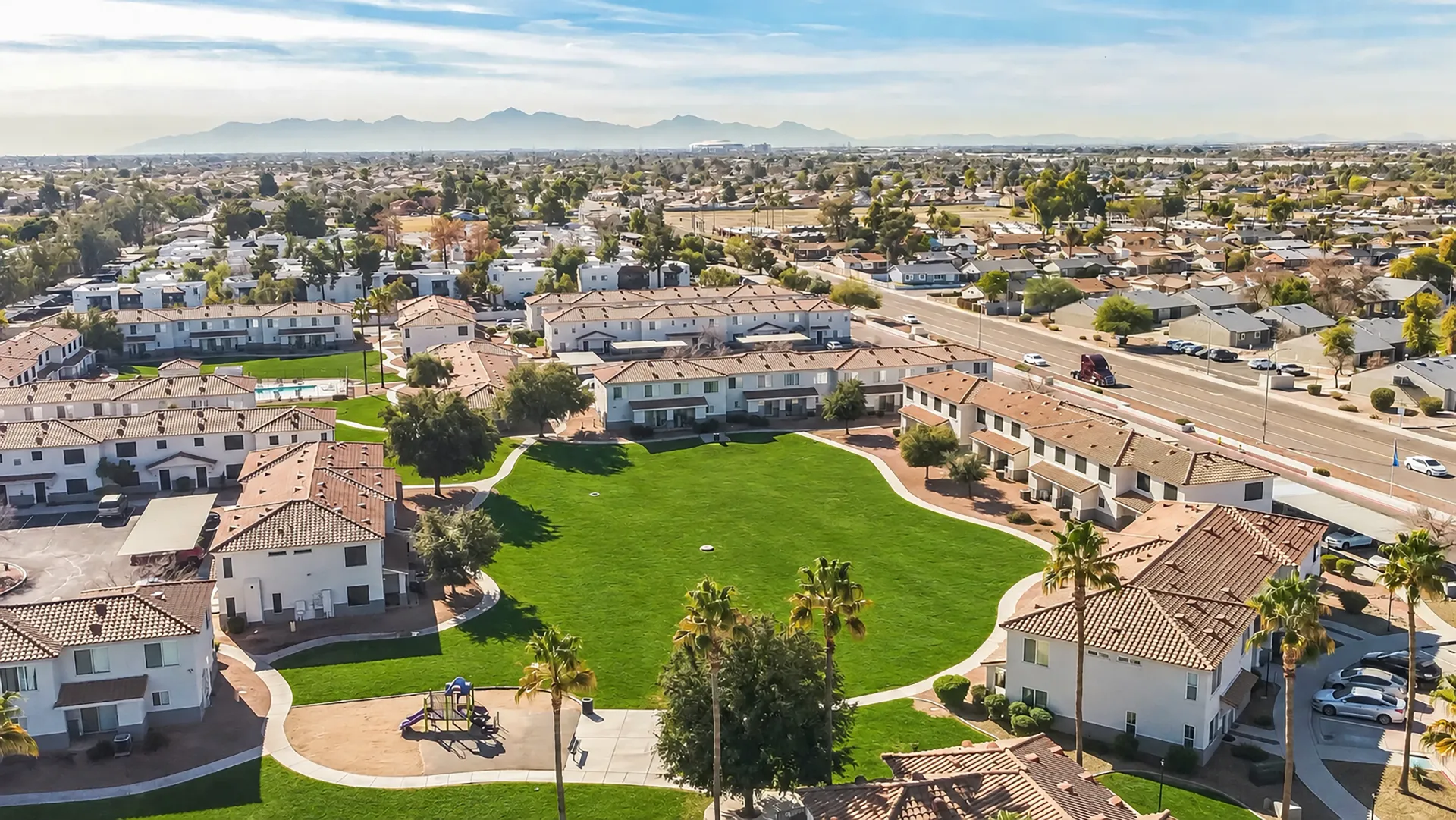 Aerial view of Rancho Del Sol courtyard with a large green lawn, walking paths, playground, palm trees, and surrounding apartment homes.
