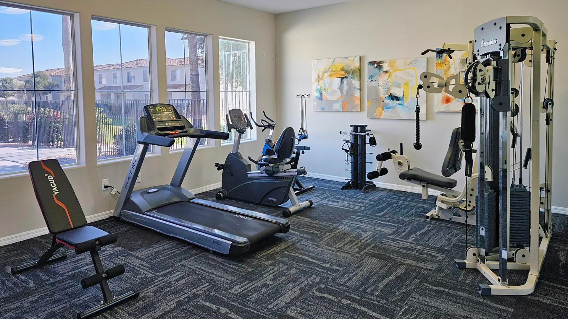 Rancho Del Sol fitness center with treadmill, stationary bike, weight-training machines, workout bench, and large windows.