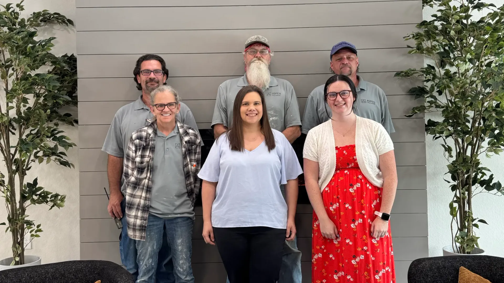 Six team members at The Avenue Apartments posing together in the leasing office, standing in front of a modern accent wall with greenery.