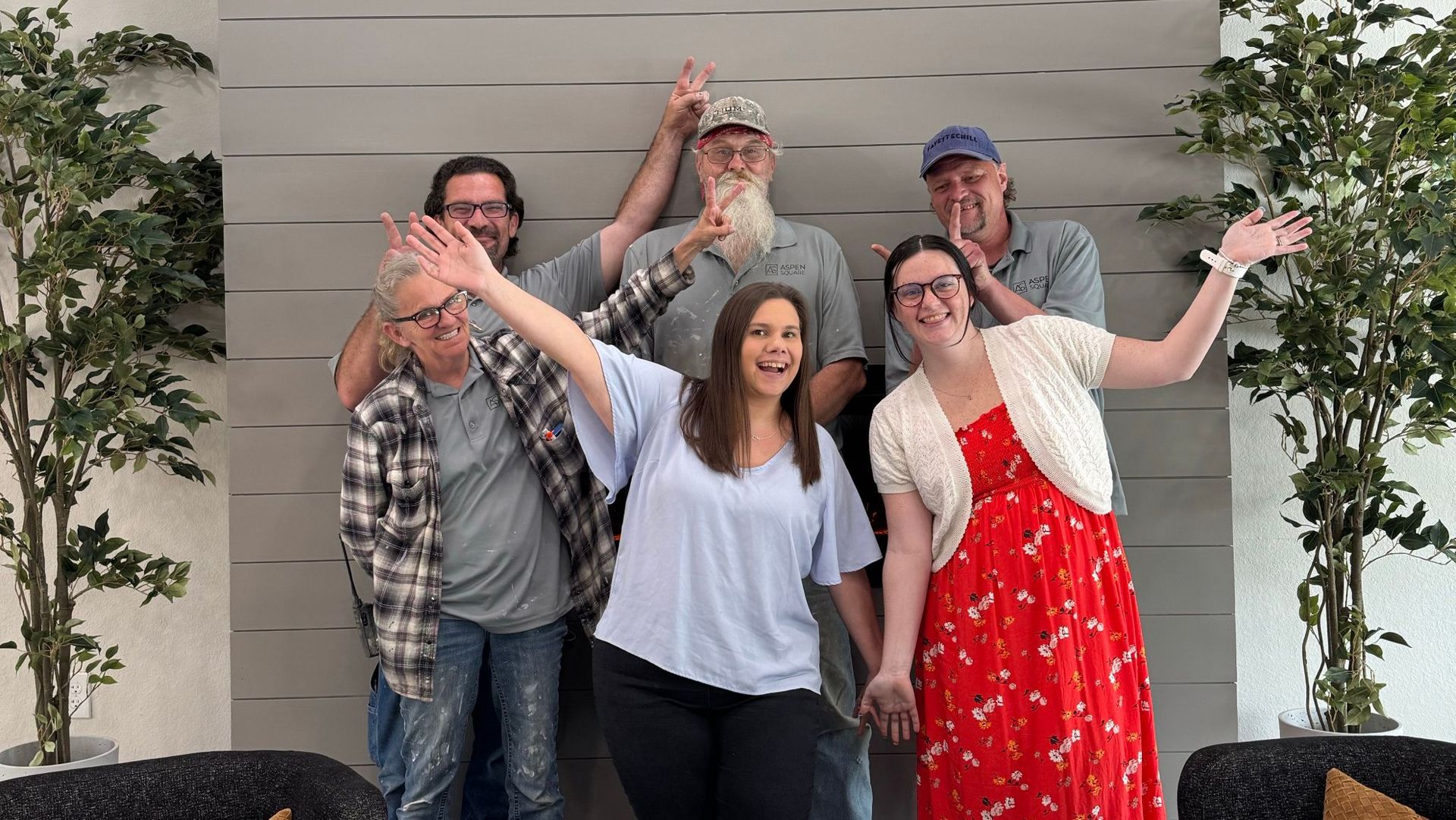 The Avenue Apartments team posing playfully inside the leasing office, smiling and making peace signs in front of a gray accent wall and modern decor.