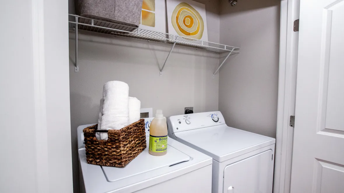 A dedicated laundry space with full-size washer and dryer appliance with overhead custom shelving for laundry supplies, ensuring a seamless and convenient laundry experience in your apartment home.