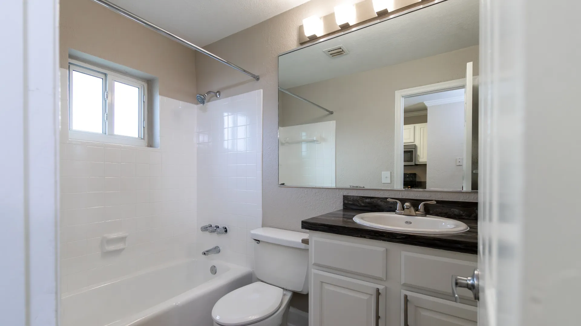 A tranquil bathroom space with a windowed shower/tub combo, white tile surround, a black fusion countertop, an expansive mirror with lighted vanity.