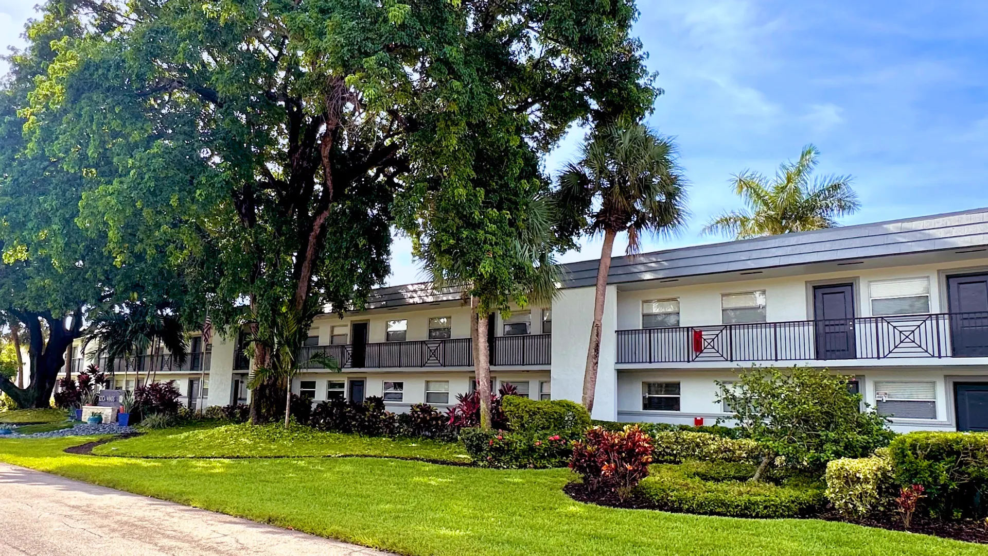 Exterior view of Boca Winds Apartments with lush green landscaping, tall trees, and well-maintained building facade.