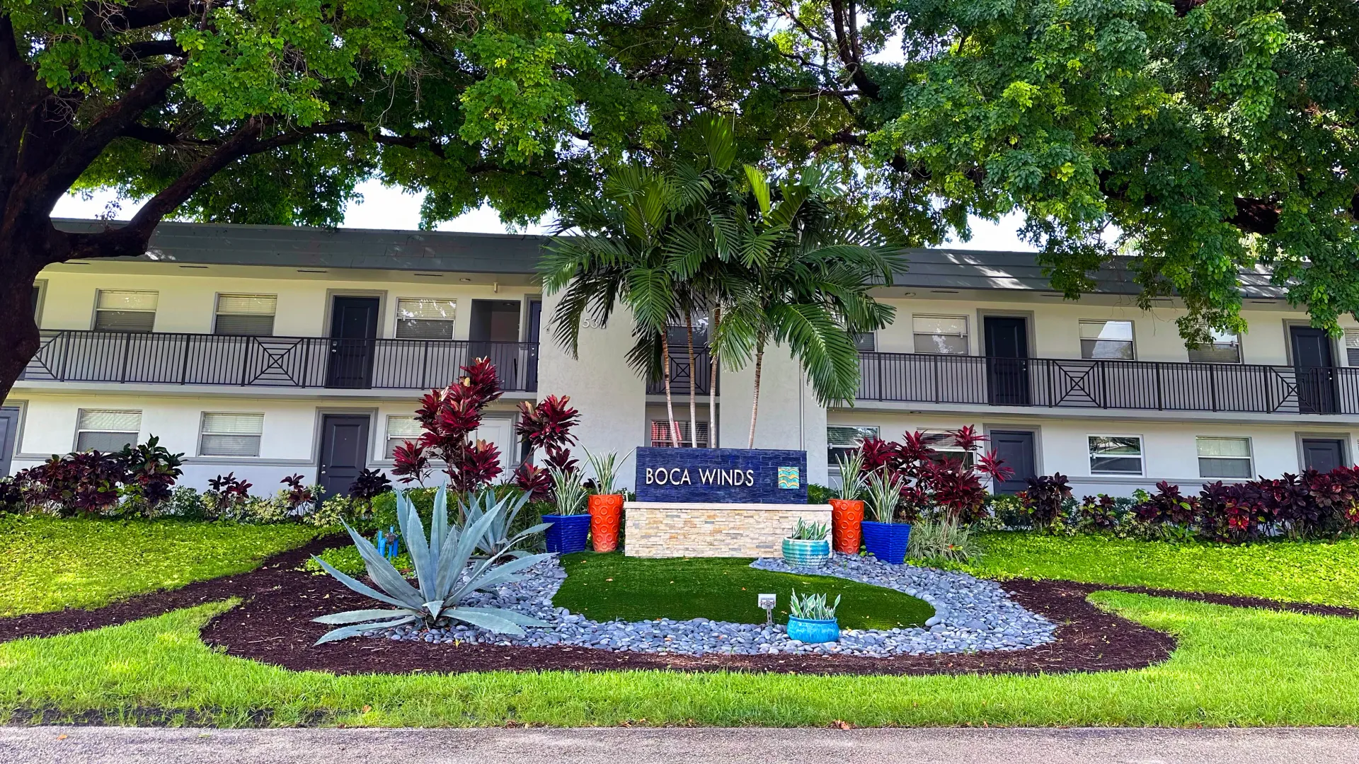 Entrance to Boca Winds Apartments with a landscaped garden, colorful planters, and a welcoming sign.