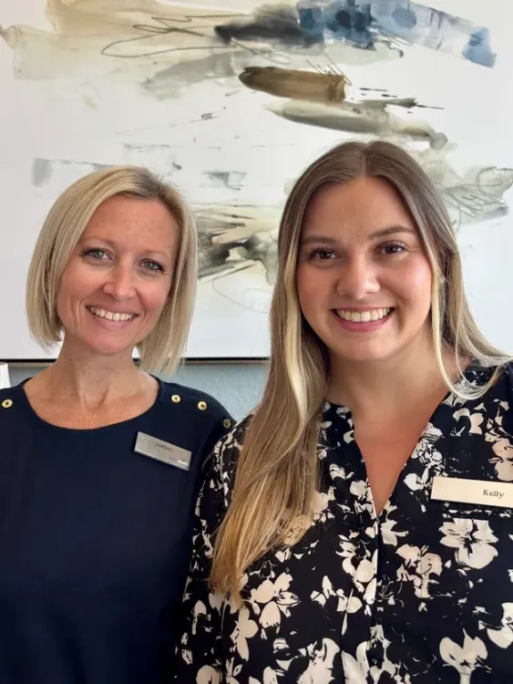 Close-up of two Eden Pointe team members smiling in front of modern wall art, dressed in black and floral tops.