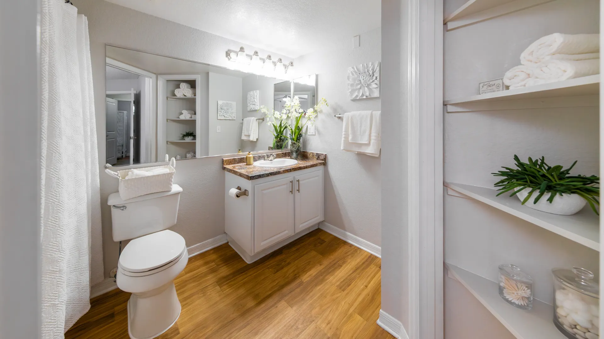Master bathroom with wood-style flooring, white vanity, large mirror, and built-in storage shelves at Ashton Chase Apartments.