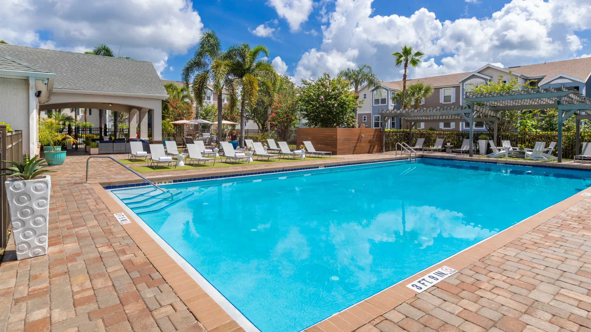 Sunny pool deck at Ashton Chase Apartments with lounge chairs, palm trees, and covered outdoor seating.