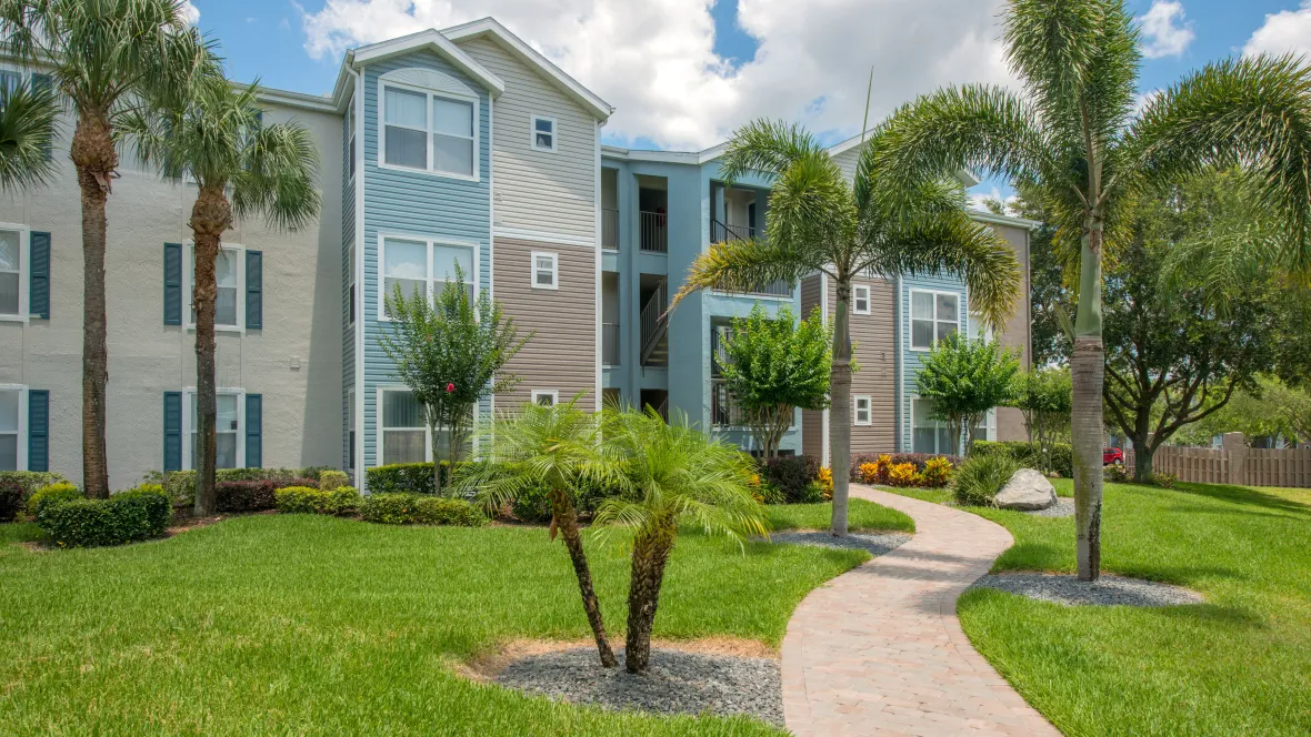 View of an Ashton Chase apartment building exterior with paved walkway, green lawn, and palm trees around the property.