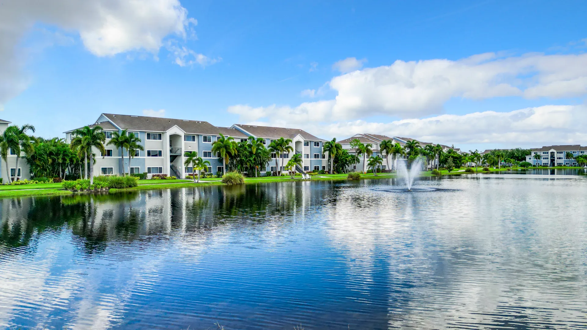 Apartment buildings and palm trees reflected in a tranquil lake with a fountain.