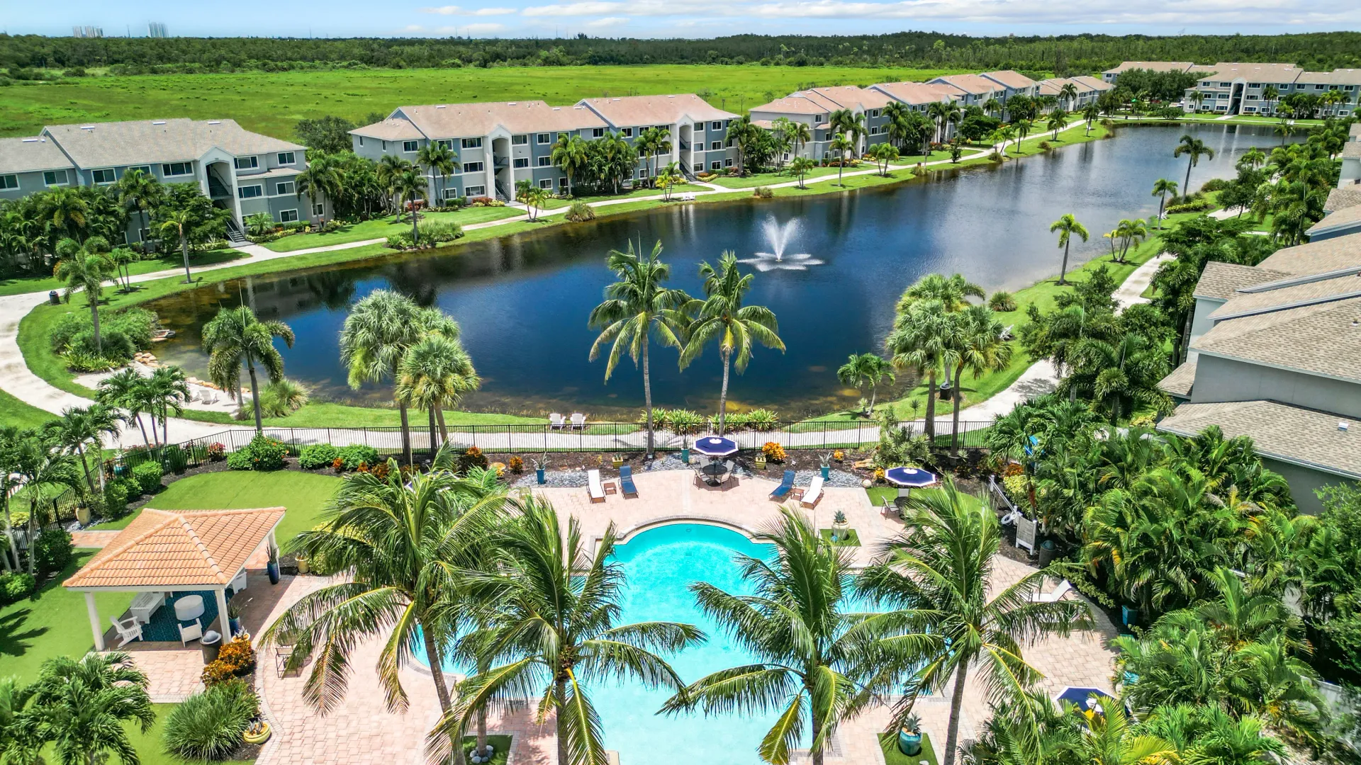 Aerial view of Lexington Palms Apartment Homes featuring a large pool, palm trees, a lake with a fountain, and well-maintained landscaping, showcasing a luxurious and serene living environment.