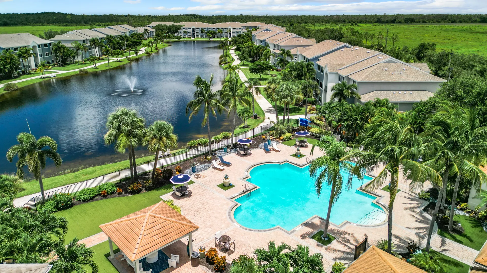 Aerial view of Lexington Palms Apartment Homes showcasing a lake with a fountain, a large pool area, and surrounding buildings, all set in lush, well-maintained landscaping.