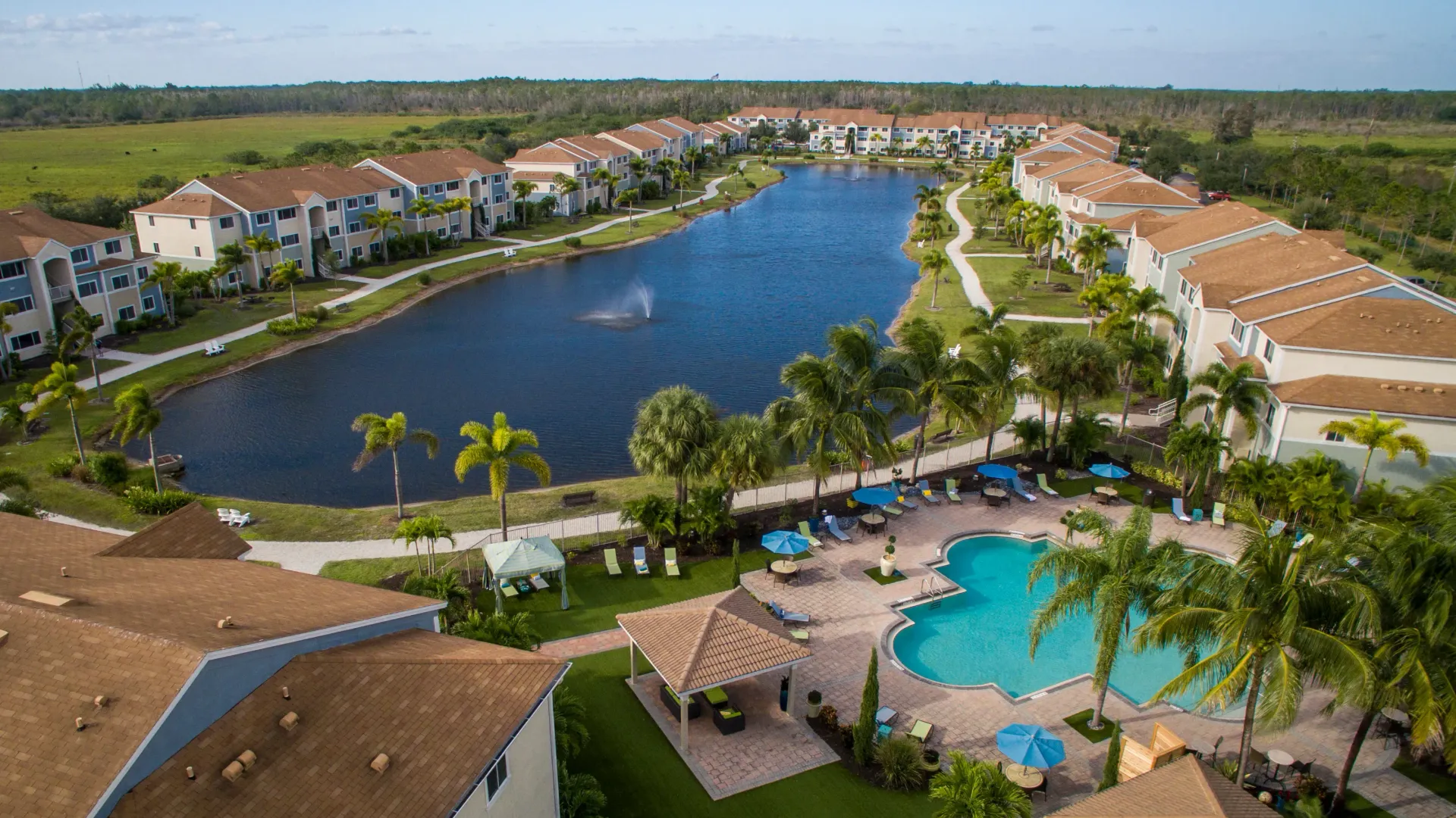 An aerial view of the community, showcasing charming lakeside apartments, lush palm trees, and Adirondack chairs by the water.