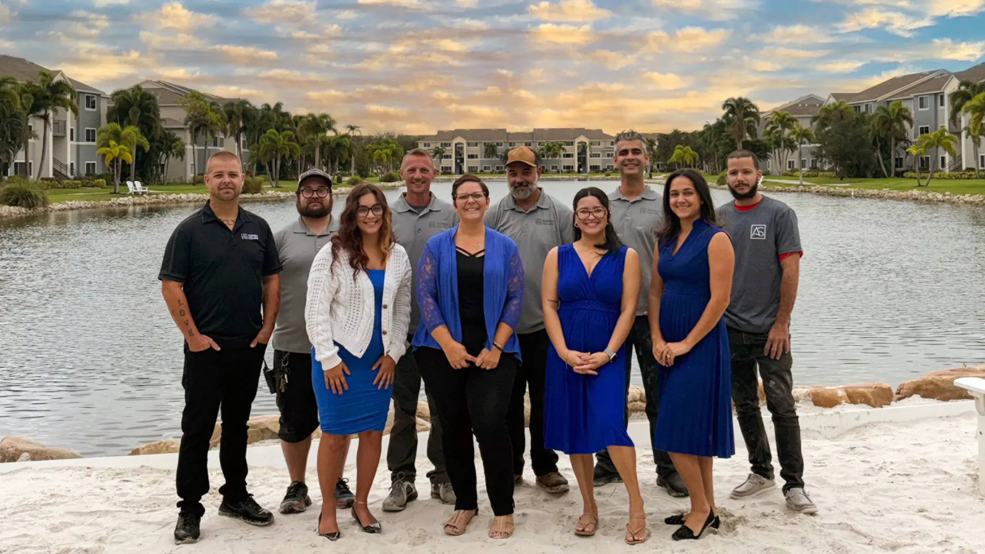 Team photo of the staff at Lexington Palms at the Forum in Fort Myers, FL, standing lakeside with apartment buildings in the background.