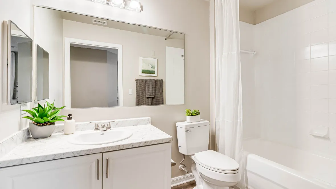 A stunning bathroom with granite-style countertops, an expansive mirror, elegant vanity lighting, and an oversized shower/tub combo with a white wrapped tile surround. 