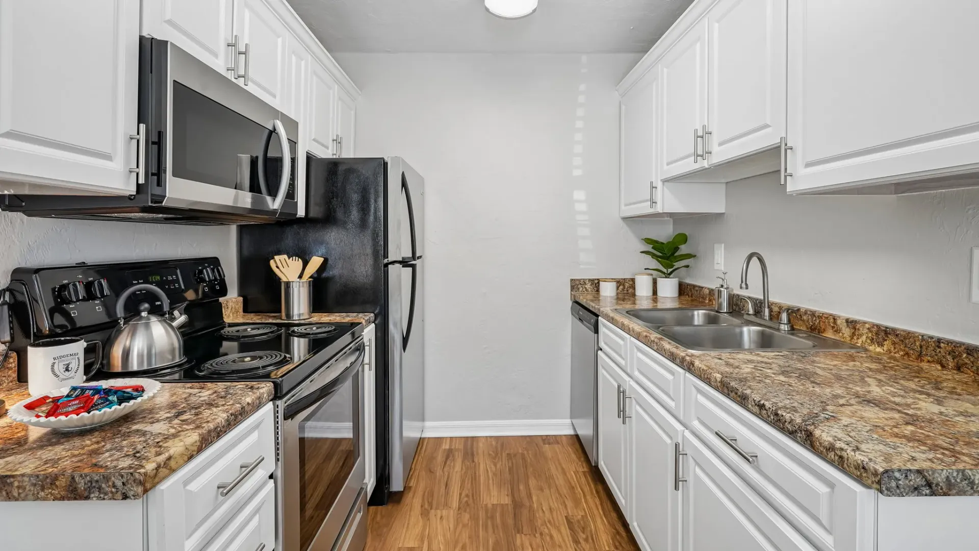 Galley-style kitchen featuring white shaker-style cabinets, granite-inspired countertops, stainless-steel appliances, and wood-style flooring.