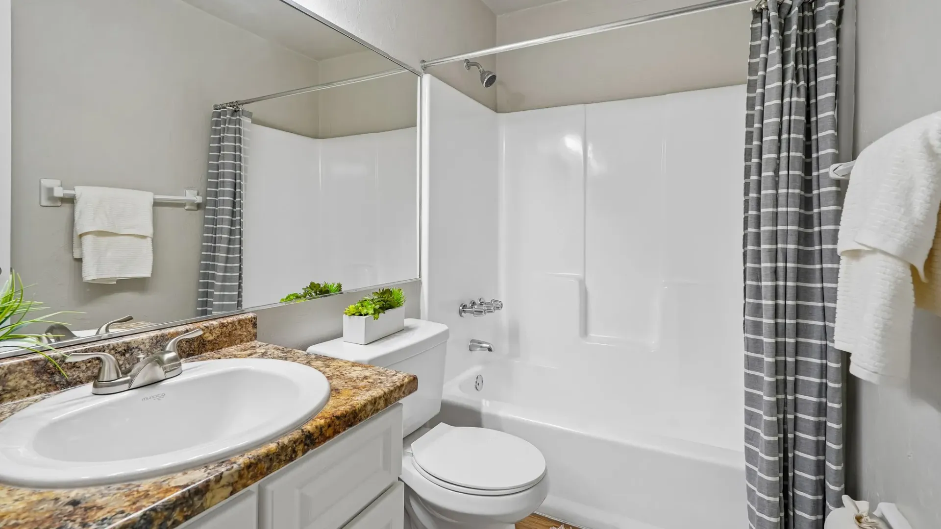 Bathroom interior at Ridgemar Commons featuring a white vanity cabinet, granite-look countertop, large wall mirror, bathtub with shower surround, and neutral finishes.