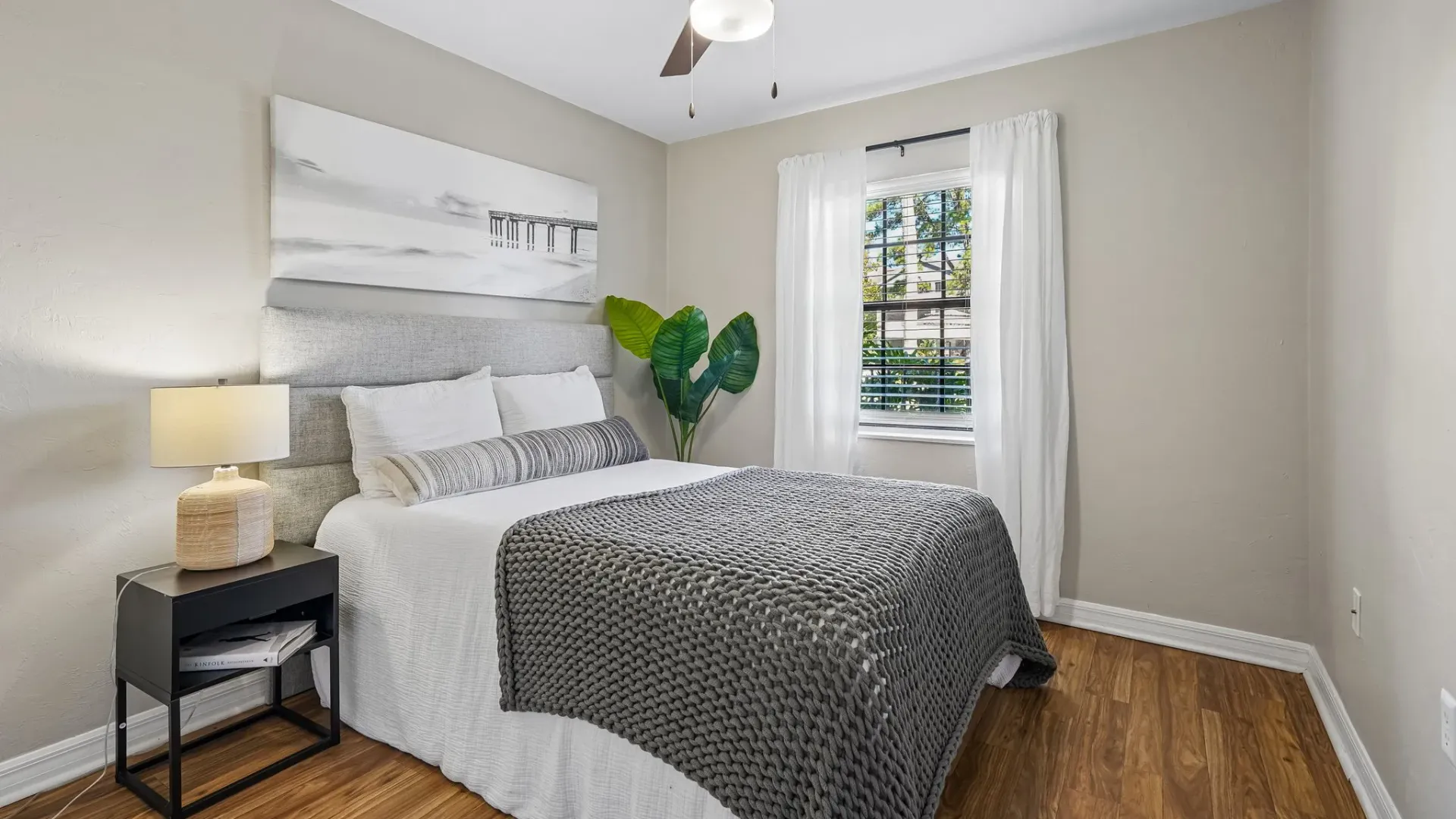 Spacious bedroom at Ridgemar Commons featuring wood-style flooring, a ceiling fan, a window with white curtains, and modern contemporary furnishings.