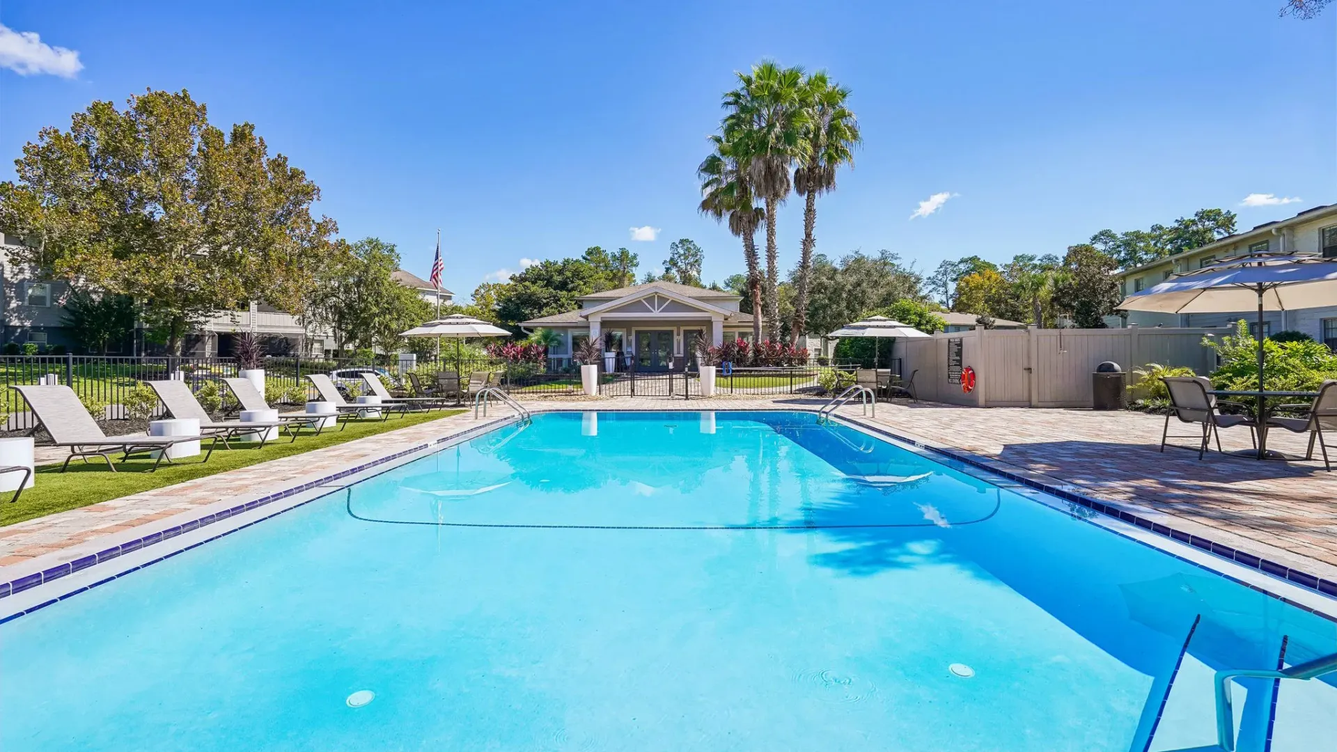 Resort-style swimming pool at Ridgemar Commons featuring crystal-clear water, lounge chairs, poolside umbrellas, palm trees, and a landscaped sundeck.
