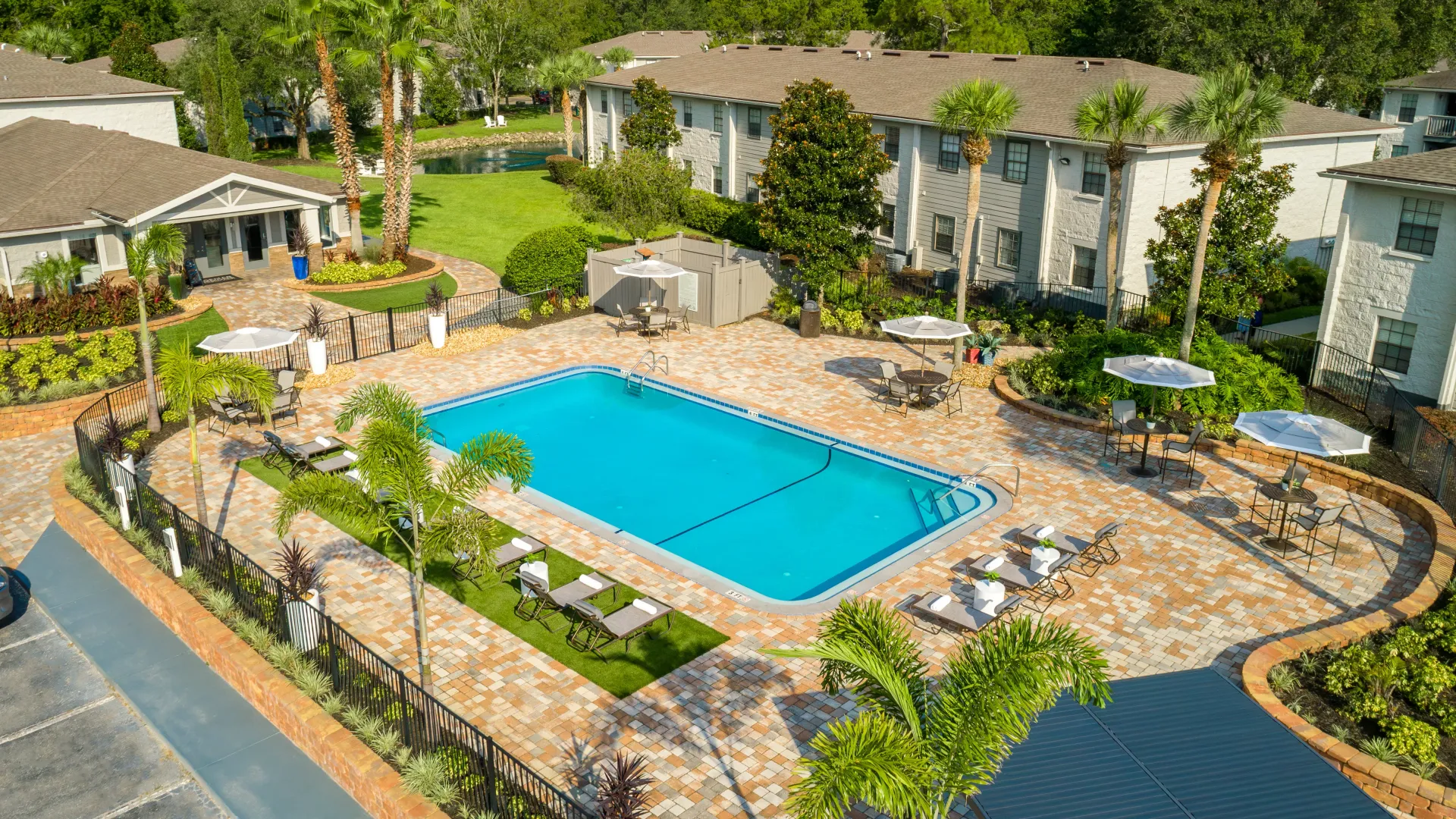 An enchanting bird’s eye view of a paradise-like sundeck with rows of poolside loungers and umbrella-covered tables, set against a backdrop of tropical palm trees and elegant pavers stretching from around the pool and leading to the leasing center.
