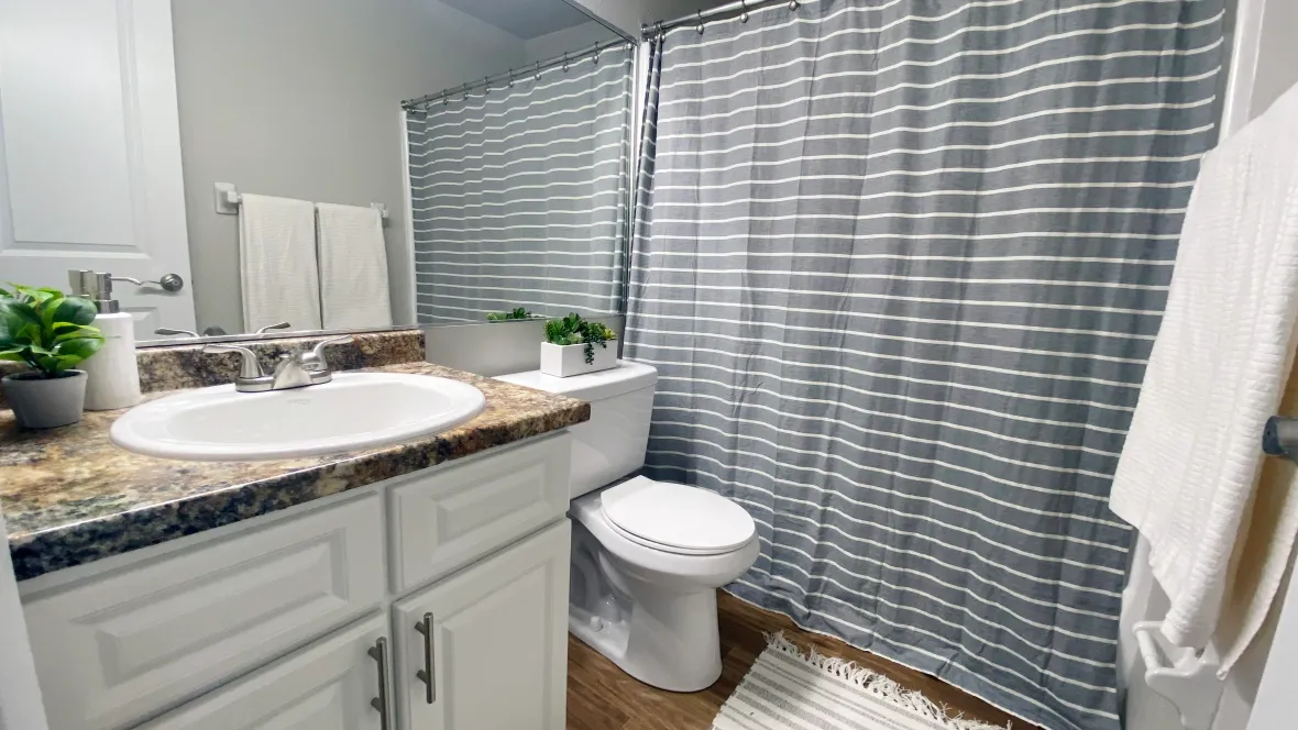 A spacious bathroom featuring a shower/tub combo, a large mirror, granite-inspired countertops, and wood-style flooring.