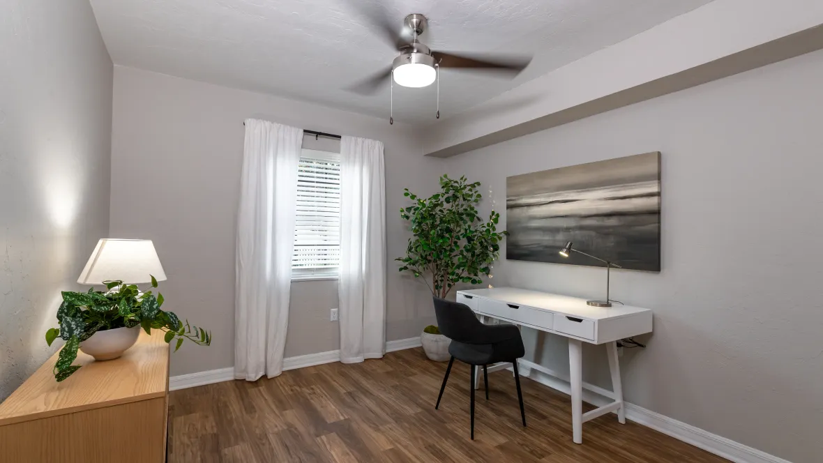 A bedroom converted into a cozy office space with stylish wood-like flooring and a modern, lighted ceiling fan, offering a perfect location for working from home or studying.