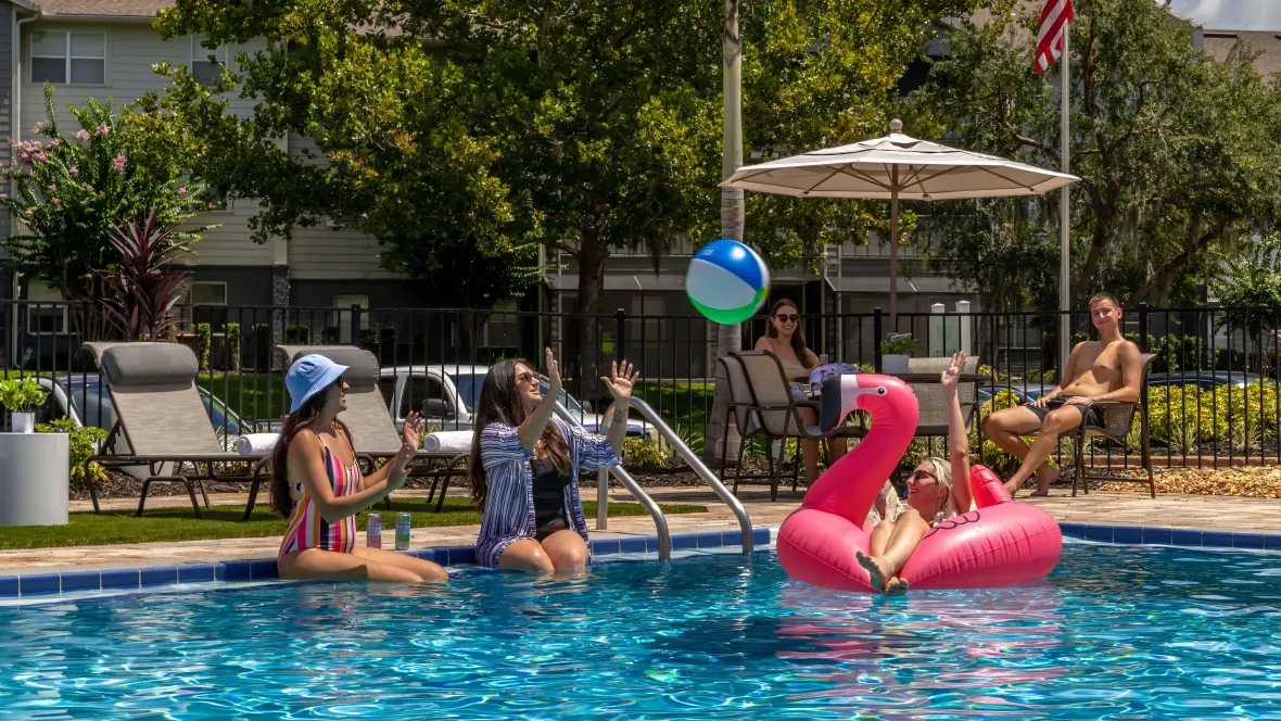 Three ladies with big smiles enjoying the glittering pool waters with a beach ball and pink flamingo float while two friends observe under a shaded umbrella with captivating smiles – all enjoying the vibrant poolside experience.