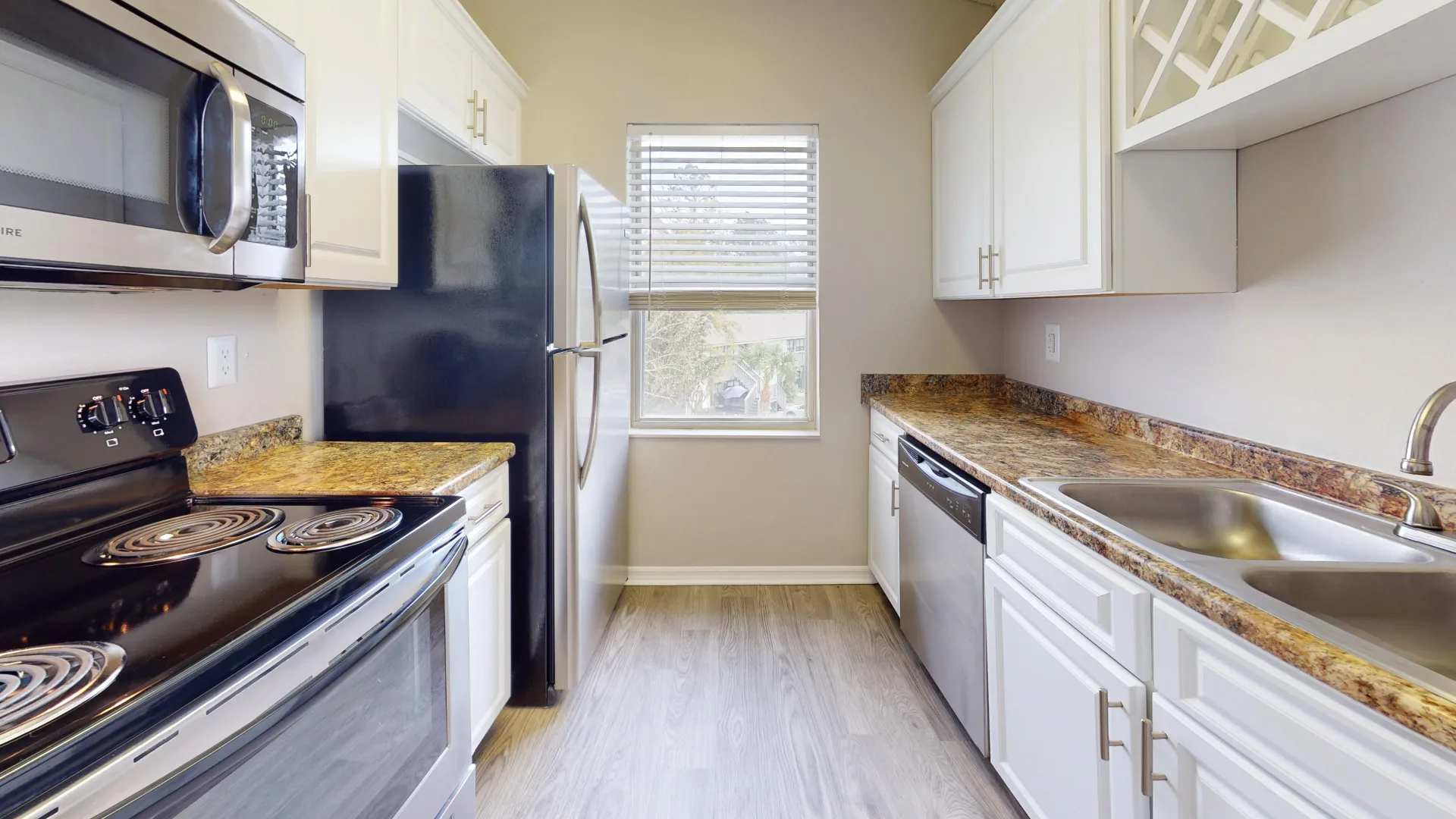 Galley-style kitchen in The City floor plan at Ridgemar Commons featuring white shaker-style cabinetry, granite-inspired countertops, stainless steel appliances, and wood-style flooring.