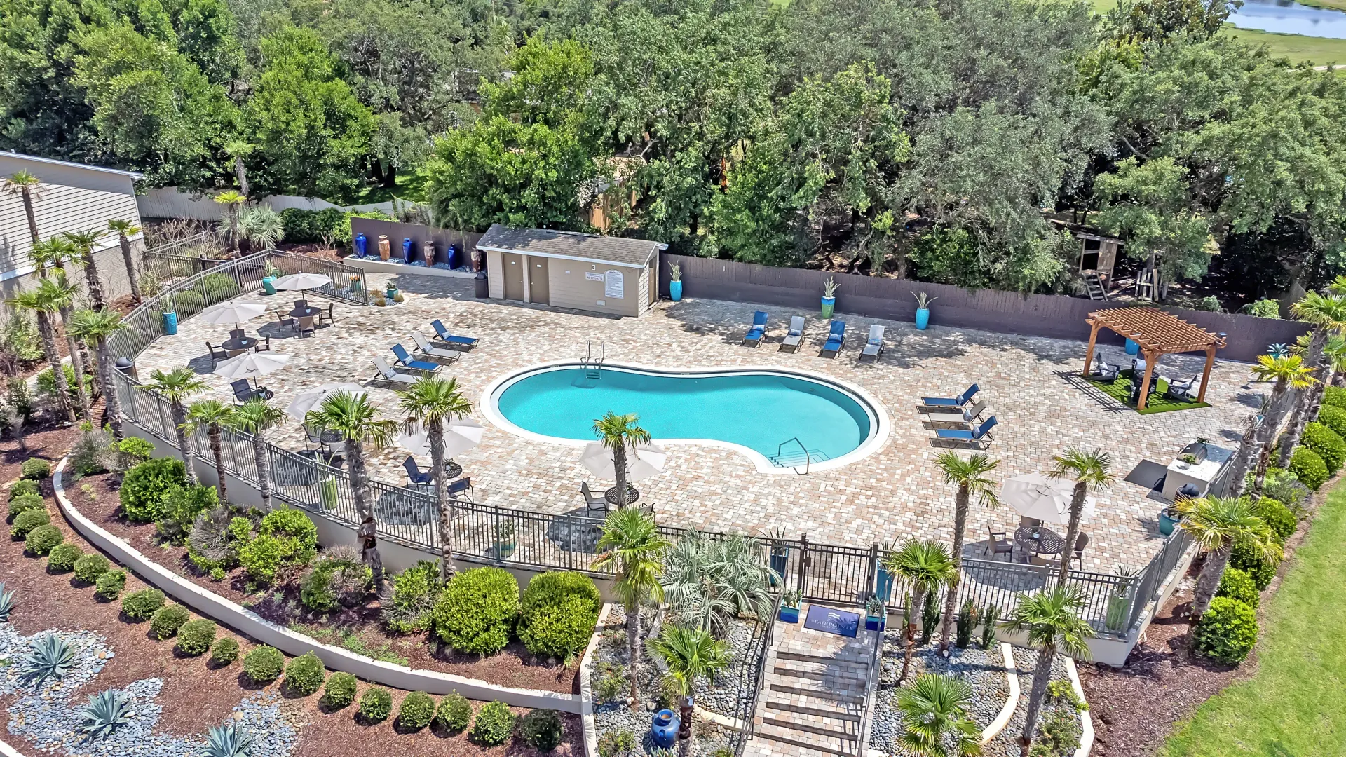 Aerial view showcasing the sparkling swimming pool, plentiful loungers and umbrella-covered tables, and lush landscaping surrounding an extravagantly paved pool deck.