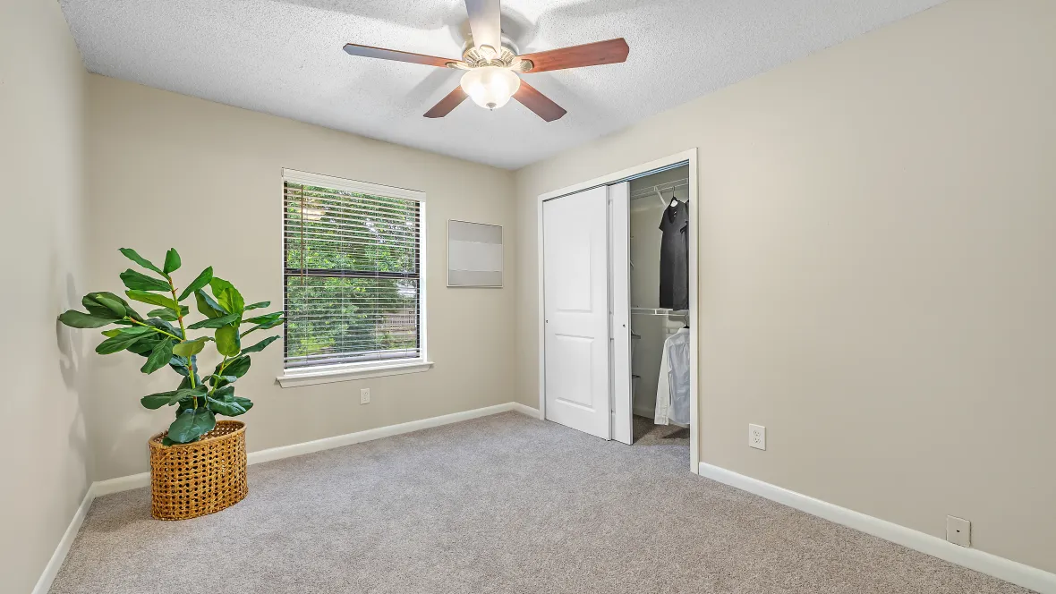A luxe carpeted bedroom boasting a wide window with privacy blinds, a capacious closet with organized shelving, and a ceiling fan with lights, promising peaceful slumber in an equipped space. 