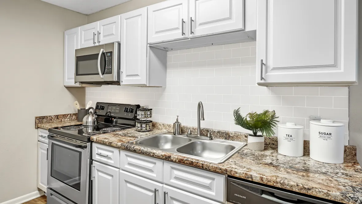 A stunning kitchen boasting extensive granite-inspired countertops, elegant white cabinetry, and a sleek muted grey subway tile backsplash that rises from counters to cabinets behind a charming double sink with a goose neck faucet. 