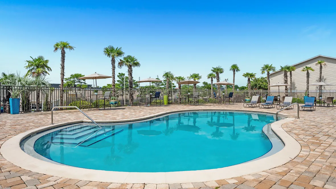  A captivating view of our expansive sundeck encircling the glistening pool, scattered by loungers and umbrella covered tables, with tall palm trees creating a picturesque skyline beyond the pool gates.