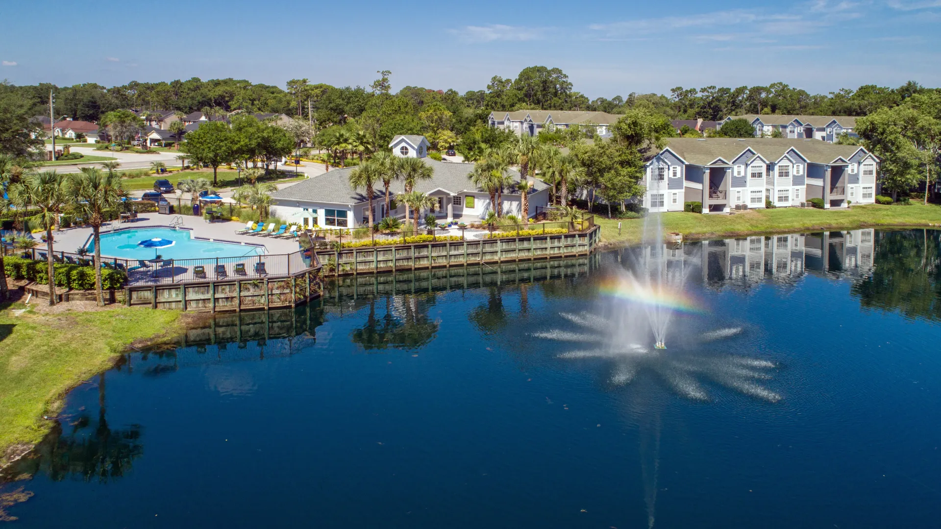 An aerial view of the community, showcasing apartment buildings, tall palm trees, the expansive sundeck, the shimmering pool, and a captivating lake with a spouting fountain highlighted by a perfectly formed rainbow.