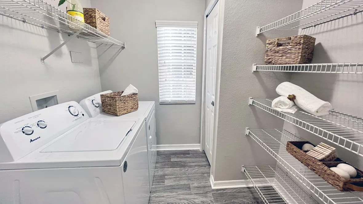 A spacious laundry room with full-size appliances, overhead shelving, versatile pantry or linen storage shelves, and a window that bathes the room in brilliant natural light.