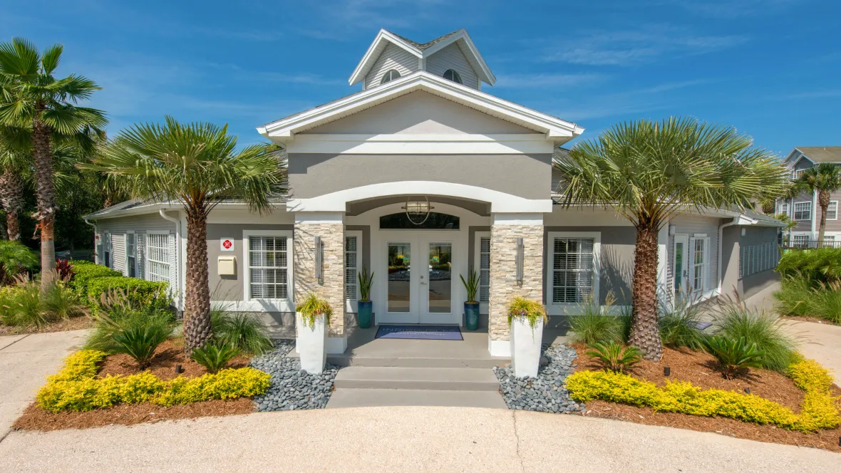 An exterior view of the leasing center's entry, framed by lush landscaping with vibrant yellow flowers and graceful palm trees on each side.