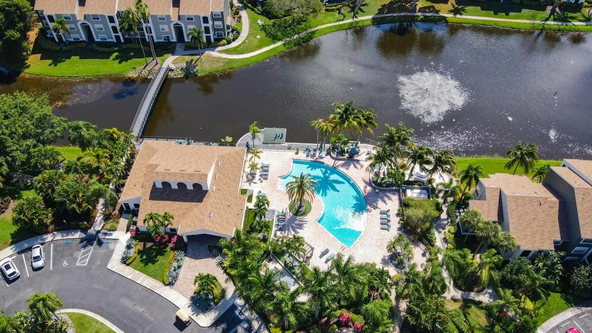 A soaring overhead community view featuring the exquisite, massive sun deck with a sparkling, half-moon shaped pool and a bridge that stretches from one side of the community to another across the lake.