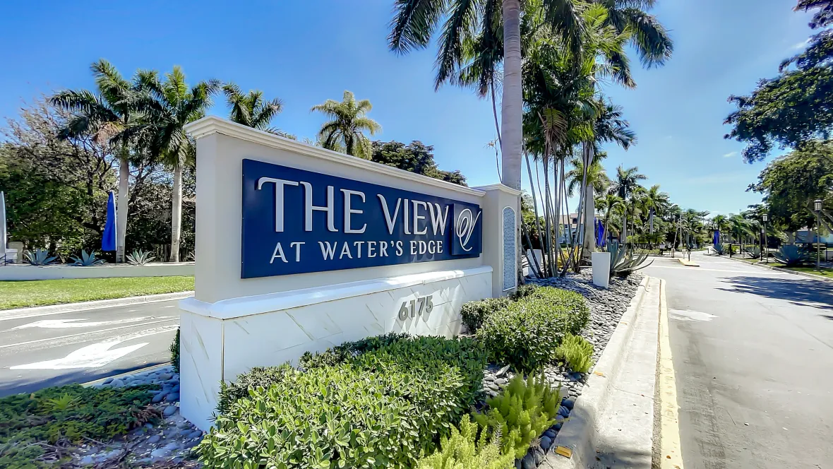 Front monument sign at the entrance drive of The View at Water’s Edge Apartments, with white lettering on a bold blue background, surrounded by lush greenery and swaying palm trees, creating a welcoming atmosphere for residents and visitors alike.