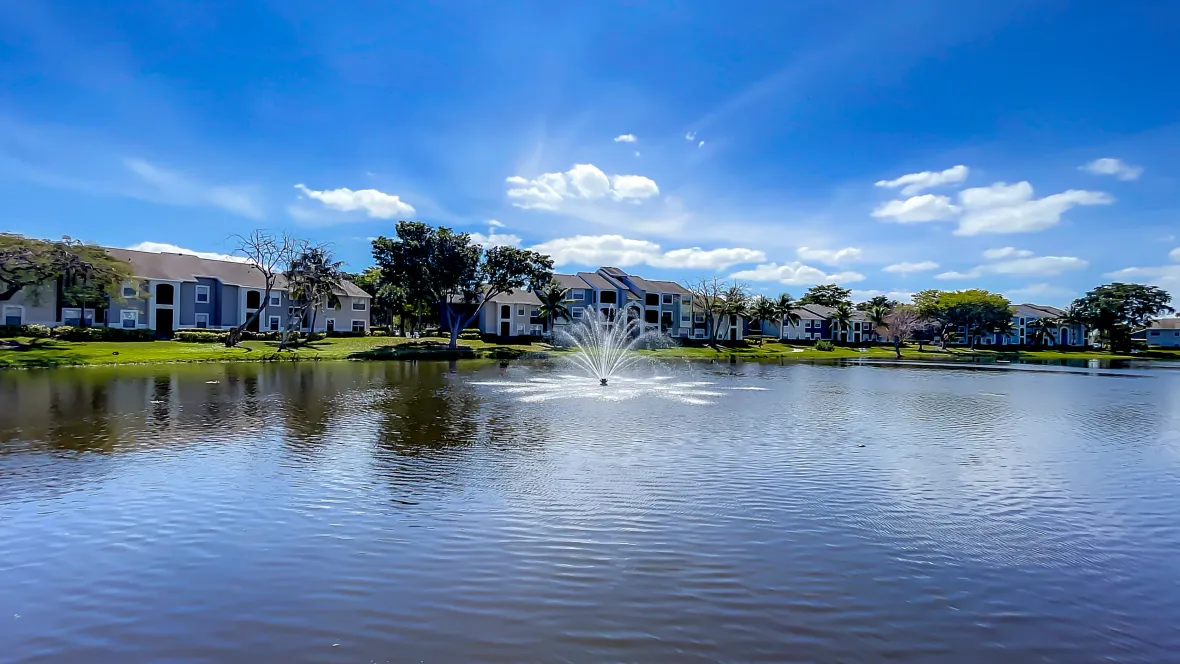 A tranquil scene overlooking Lake Osborne, featuring glistening waters and a soothing fountain next to well-maintained apartment buildings, surrounded by lush greenspace—a true lakeside sanctuary.