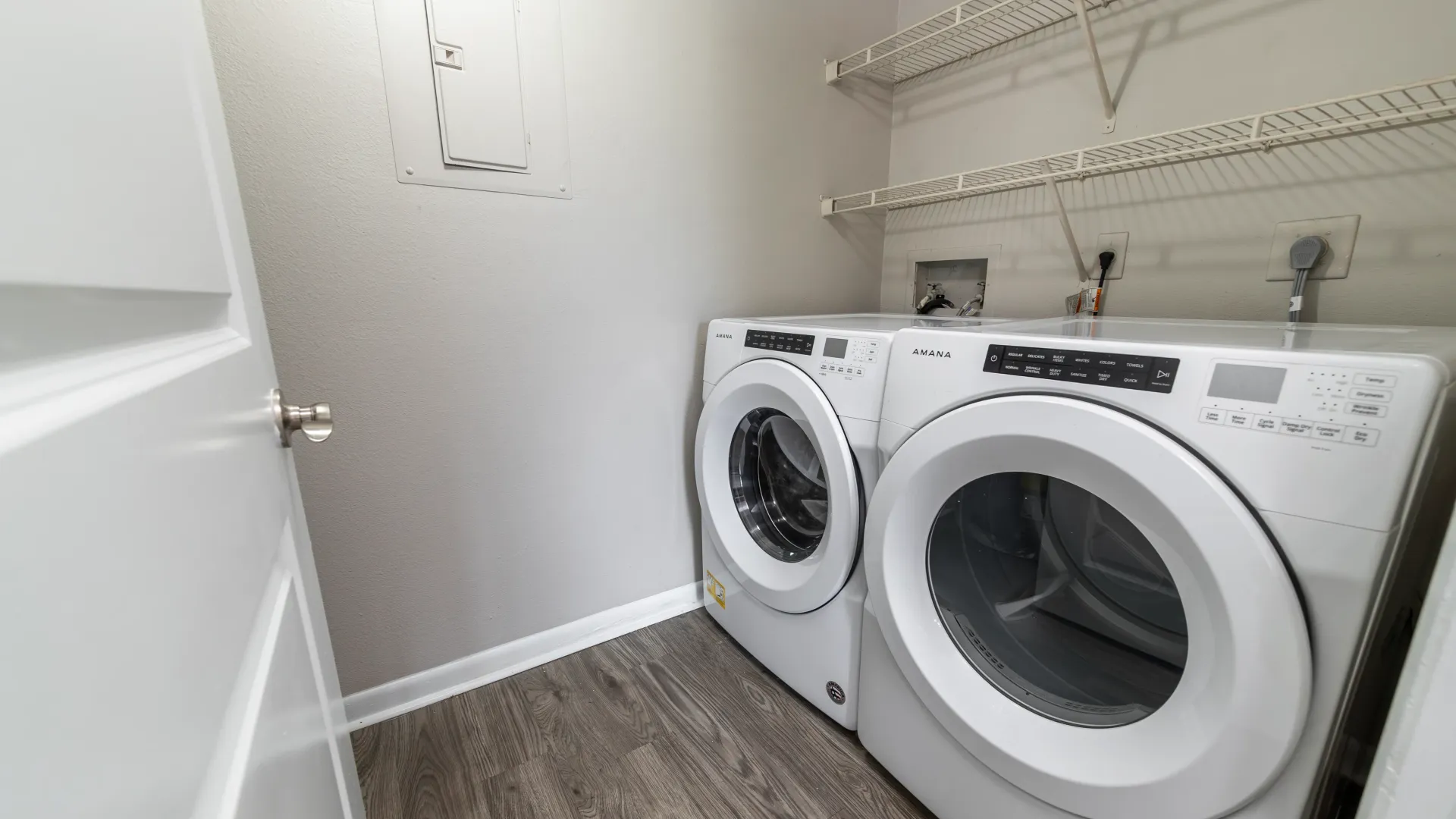 An accessible laundry room just off the kitchen showcasing a washer and dryer with front-loading convenience and two rows of overhead open-wire shelving. 
