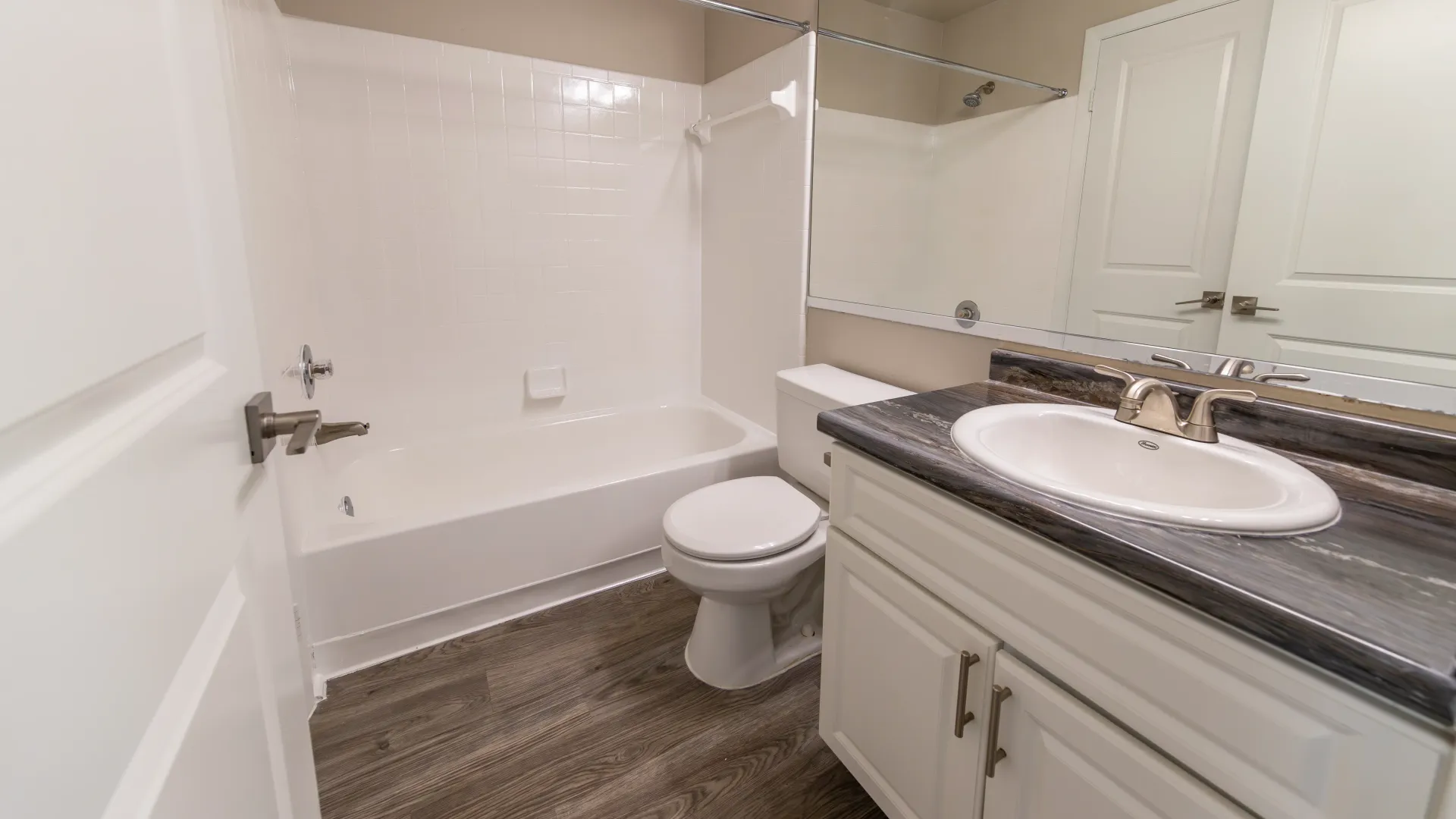 A magnificent bathroom adorned with black fusion countertops, a sprawling mirror, wood-inspired flooring, and sleek under sink cabinetry.