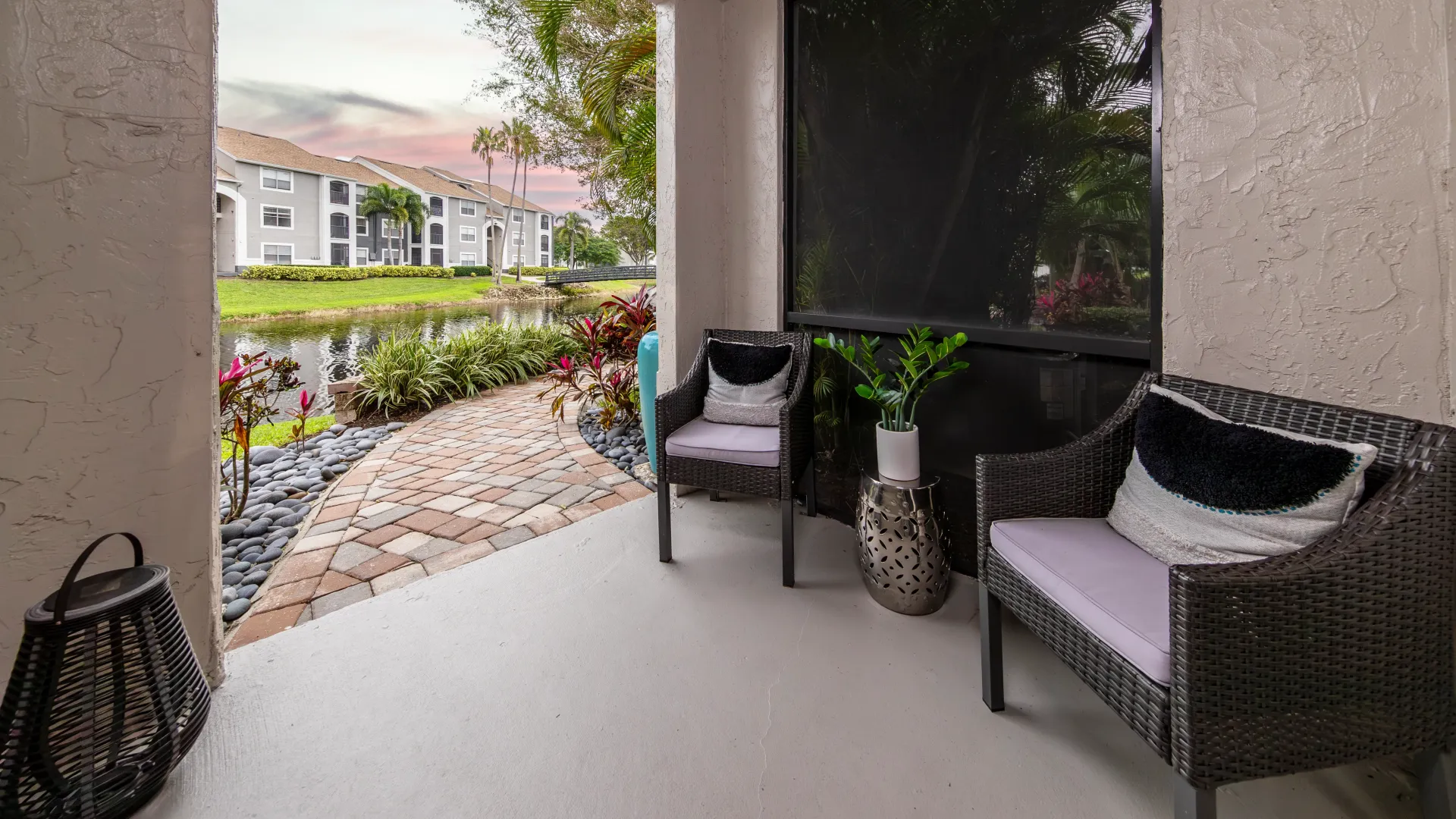 A welcoming paved walkway leads directly to a screened-in patio on the ground floor staged with two wicker chairs and a decorative side table for a cozy, homey sanctuary—an extension of the living room. 