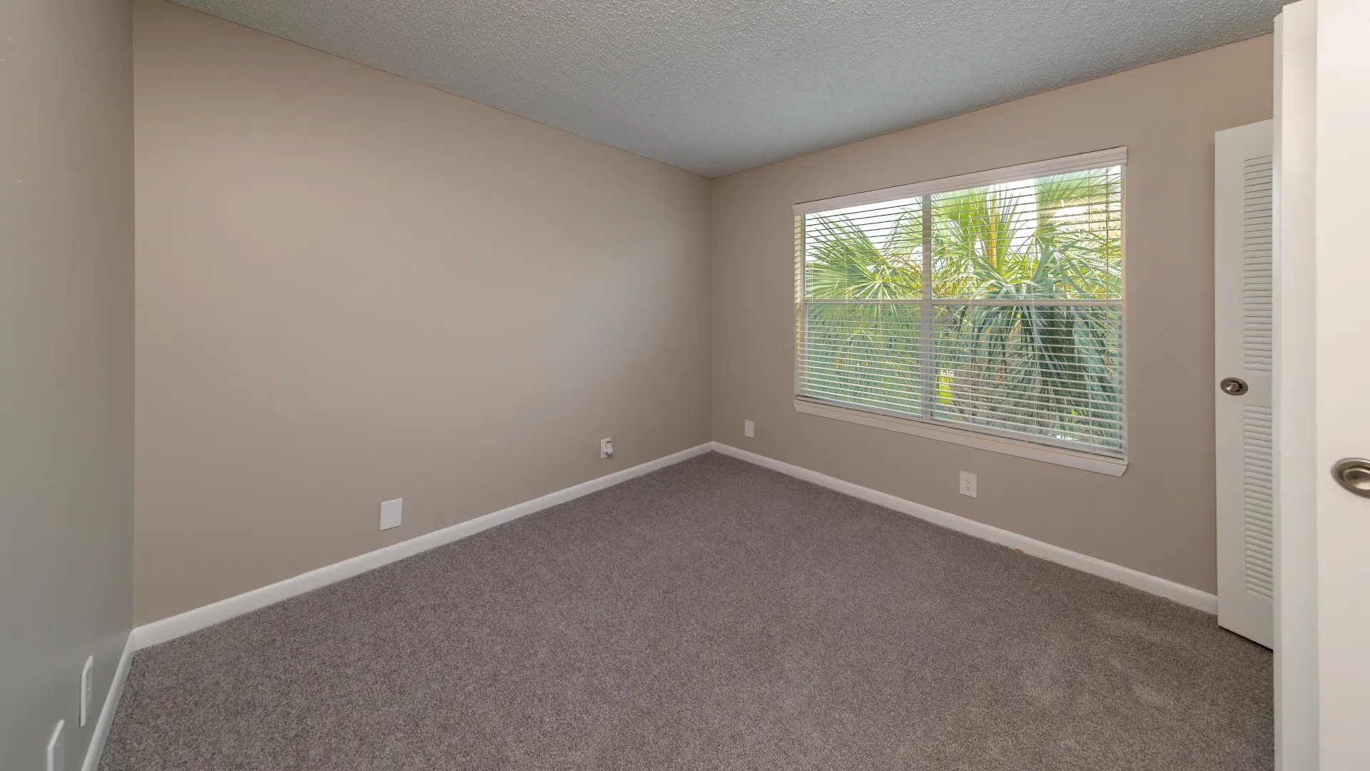 A spacious guest bedroom, adorned with brilliant lighting streaming through large windows outfitted with wood-style blinds, complementing neutral-colored plush carpeting. 